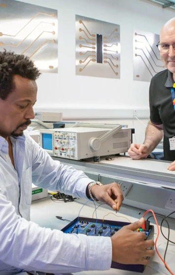 A lecturer and a student in an electronics lab working on a circuit board experiment. The lecturer is smiling while overseeing the student’s work. A lecturer and a student in an electronics lab working on a circuit board experiment. The lecturer is smiling while overseeing the student’s work.